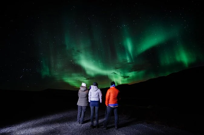 Drei Menschen unter grünen Nordlichtern über einer dunklen Landschaft